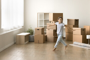 Happy active kid girl running past stacked moving boxes in empty room, dancing, hopping,...