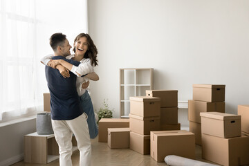 Happy married couple enjoying first relocation, moving activities, hugging, having fun at heap of transportation cardboard boxes. Strong husband embracing and lifting laughing wife
