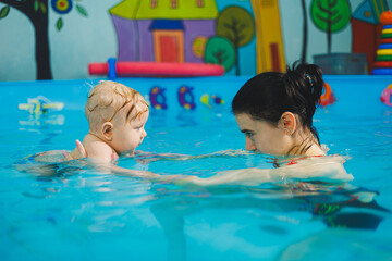 Obraz na płótnie Canvas A female trainer teaches a baby to swim in the pool and supports his hands. Baby swimming in the pool. Teaching a newborn boy to swim in the pool