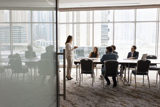 Wide shot of multiethnic business team talking at meeting table in office conference room, brainstorming in co-working space with large window. Project leader woman talking to colleagues