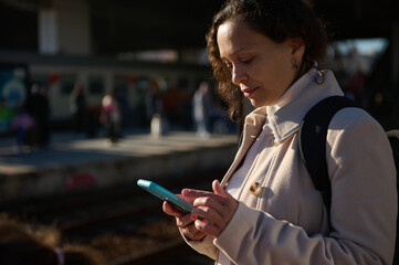 Young woman standing on the train station railway platform, checking her mobile phone white waiting to board the train - obrazy, fototapety, plakaty