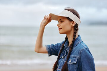Joyful Summer Escape: A Beautiful Young Woman Enjoying the Sun and Sea in a Casual Fashion Dress, Hat, and Sunglasses, Standing on a White Sandy Beach with Blue Ocean and Sunny Sky in the Background.