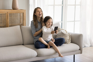 Obraz premium Little pretty daughter sits on laughing loving mothers laps, holds smartphone, having fun, make photos, play new game, enjoy mobile application, talk to friend using videocall app on modern cellphone