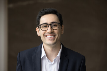 Happy Arab business man in formal office jacket and glasses looking at camera with toothy smile, laughing, posing indoors for front head shot. Young handsome businessman, employee video call portrait
