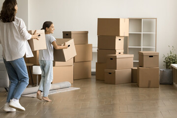 Fototapeta premium Pretty daughter kid and young mom carrying moving boxes in new apartment, walking to heap of stacked cardboard containers, enjoying relocation activity after buying house