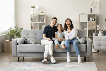 Joyful young parents and sweet kid girl sitting on soft comfortable grey couch in cozy home...