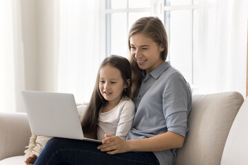 Fototapeta premium Mother and little daughter spend weekend leisure on internet use laptop, sit on sofa look at device screen, watch educational program, buy in marketplace, enjoy e-commerce services and cartoons online