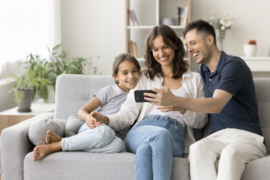 Happy young parents and cute daughter kid watching content on smartphone, using gadget for online communication, taking family selfie on cozy home couch, enjoying Internet technology