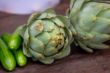 Selective focus of green raw Artichoke and cucumbers on wooden table, Fresh vegetable in market stall, The globe french artichoke is a variety of a species of thistle cultivated as a food.