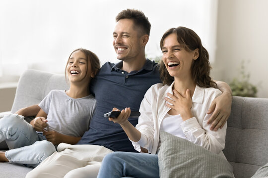Cheerful excited young mom, dad and daughter kid watching TV at home together, relaxing on couch, using distant remote control, looking away, laughing, enjoying movie, series, Internet television