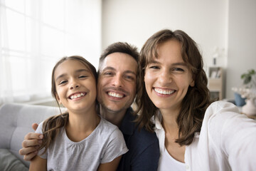 Happy young parents and cute girl kid looking at camera with toothy smiles, posing for home family...