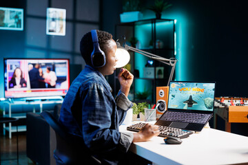 African American female gamer competes in an online video game, using laptop and wireless headset...