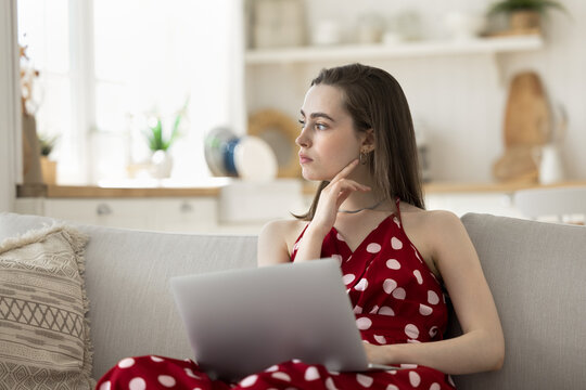 Pensive pretty young woman sit on couch with laptop, deep in thoughts, think on answer, make college assignment, faced up with difficult task, spend time on internet, doing telework, chatting online