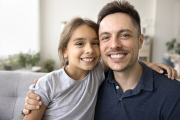 Happy loving daddy holding cheerful cute daughter kid in arms, looking at camera with toothy smile. Positive young Father and adorable child hugging, sitting close on home couch, posing for portrait