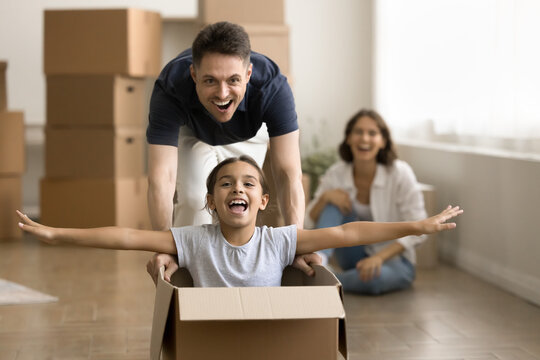 Excited Strong Young Dad Walking And Driving Relocation Box With Cheerful Kid Inside, Playing With Daughter. Family Celebrating Apartment, House Buying, Having Fun Among Paper Containers