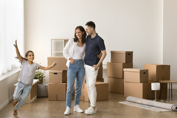 Fototapeta na wymiar Happy young family celebrating house buying, mortgage. Parents standing at heap of cardboard moving box in empty room, hugging, smiling, looking at kid running around