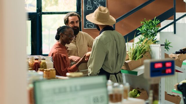 Farmer offers fresh veggies samples to couple shopping for groceries, diverse customers looking to buy potatoes and pantry supplies for healthy eating. Local eco storekeeper recommending produce.