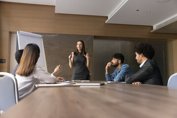 Fototapeta premium Excited project leader woman presenting ideas for startup to multiethnic team, standing at meeting table, discussing creative plan, strategy with sitting colleagues, employees