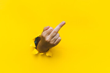 Middle finger of right hand, insulting gesture. Torn hole in yellow paper. Fuck you concept. Aggressive reaction. - obrazy, fototapety, plakaty