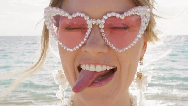 Portrait of funny hilarious woman in pink glasses making silly faces, pretending singing outloud on beach. Positive girl having good time showing tongue and playing kissy-face. Closeup emotional bride