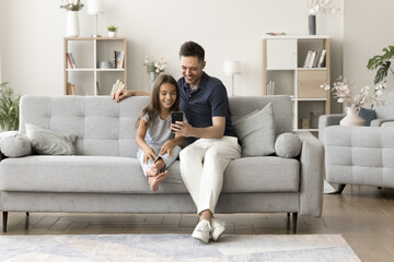Cheerful sweet daughter kid and happy young daddy using mobile phone on sofa together, talking on video call in stylish living room, enjoying online communication in cozy home interior