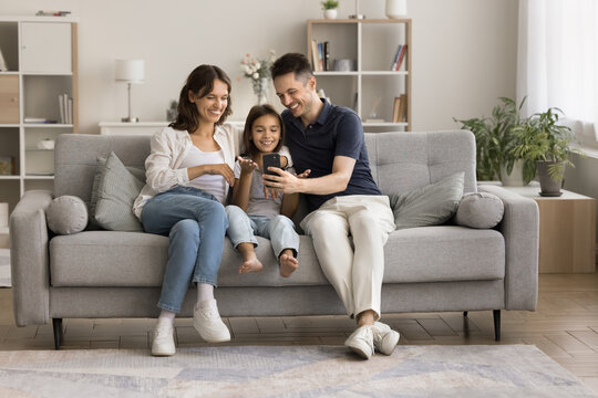 Happy young couple and little kid talking on video call, using smartphone for online family conference communication, resting on comfortable couch together, holding cell, speaking, smiling