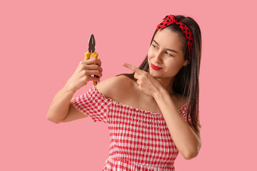 Portrait of young pin-up woman pointing at pliers on pink background. Women's History Month