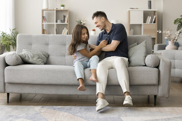Happy excited young dad tickling cute daughter kid on soft stylish couch, playing active game with child in cozy living room, cuddling girl, laughing, having fun, enjoying funny leisure