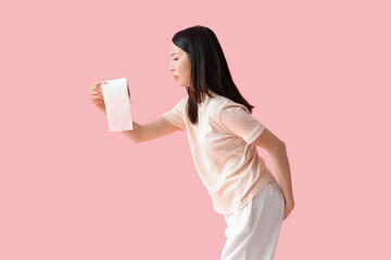 Young Asian woman with hemorrhoids and toilet paper on pink background