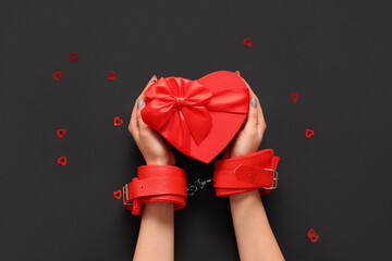 Female hands in handcuffs from sex shop and heart-shaped gift box on dark background, closeup....