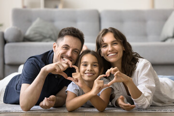 Cheerful attractive mom, dad and sweet daughter kid joining fingers, showing hand heart shapes, resting together on warm floor at cozy home, looking at camera with toothy smiles for family portrait