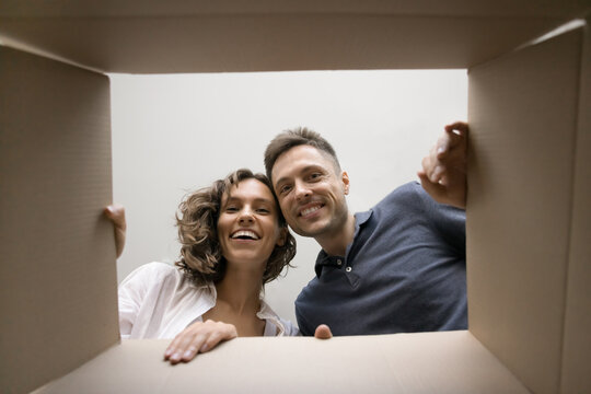 Cheerful young couple opening parcel, looking inside cardboard moving box with joy. Happy man and woman unpacking awaiting carton package from delivery service. Low angle, bottom view portrait