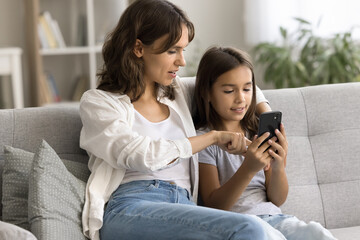 Engaged beautiful mom showing cute child girl how to use online application, service on mobile phone, resting on cozy soft home couch, touching smartphone screen, speaking, hugging kid