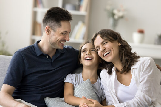 Cheerful excited parents and kid having fun together on comfortable couch, laughing, hugging, enjoying close relationship, parenthood, childhood, family leisure time at home