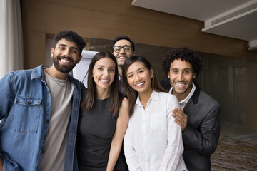 Cheerful multiethnic team of office friends standing close together, hugging looking at camera, smiling, laughing. Successful business colleagues posing for portrait, enjoying corporate friendship