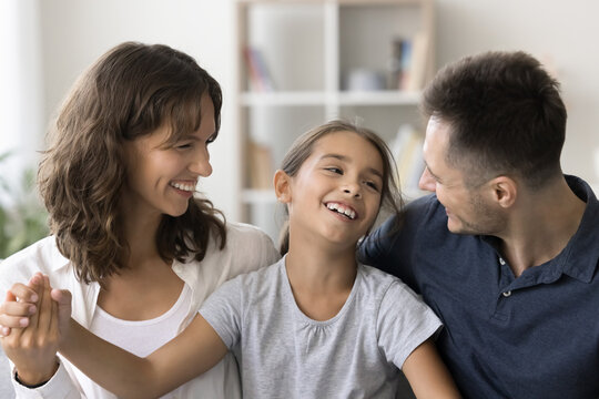 Cheerful young parents and happy carefree child girl sitting close together, talking, laughing, hugging, enjoying family leisure, relationships, childhood, parenthood. Mom and dad cuddling kid