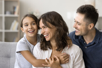 Carefree cute little girl kid hugging positive mom and dad on couch, smiling, laughing. Happy couple of parents playing with child on sofa, enjoying care, family leisure time at home