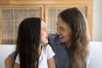 Close up shot of cheerful young mom and cute little daughter sit on sofa, hugging, looking at each other with love, smile, enjoy priceless time together at home. Happy motherhood, custody and adoption