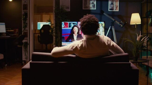 Man holding remote, looking at latest news on TV after zapping through channels. Zoom in shot on person using ultrawide smart TV in neon lit living room to entertain himself after work