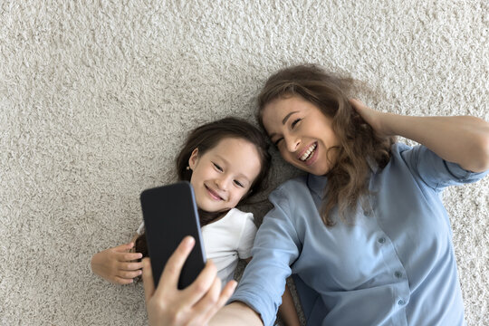 Happy mother and daughter lying on floor looking at cellphone screen, take selfie pictures, having fun using new mobile application, spend free time on internet, make videoconference call, top view