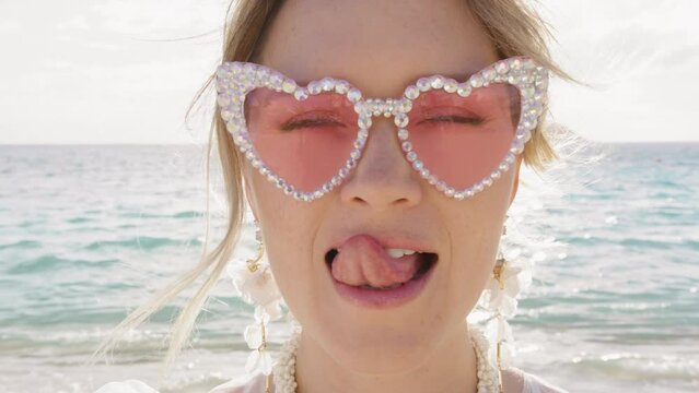 Closeup portrait comic young woman flirting with camera. Smiling girl in fancy pink glasses licking lips, taking off heart shape glasses and winking with one eye, blowing air kiss. Romantic date shot