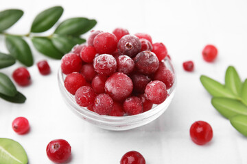 Frozen red cranberries in bowl and green leaves on white tiled table, closeup