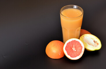 A tall glass of fruit juice on a black background, next to pieces of ripe mango and grapefruit.