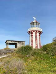 Red lighthouse with white stripes on blue sky. Coast lighthouse at the top of a green hill. Harbor in the island.	