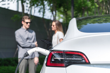 Young couple travel with EV electric car charging in green sustainable city outdoor garden in...