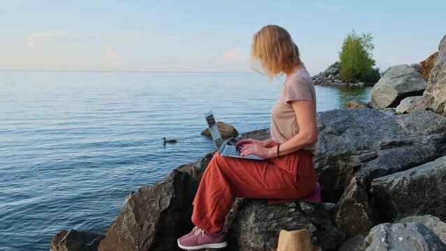 A digital nomad, blonde caucasian woman sits by the sea and works remotely. Female freelancer uses laptop computer on empty seashore. Office for introvert outdoor.