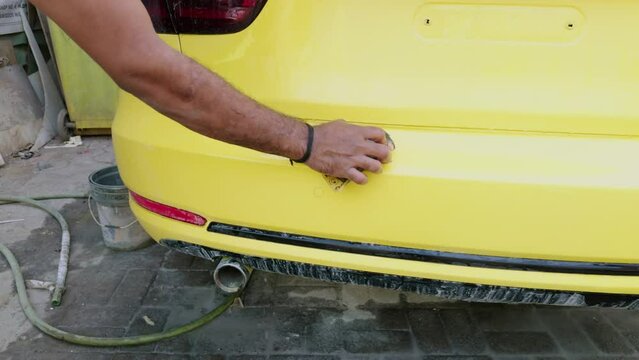 Man hand washing a yellow sports car before painting with a sponge and bucket of water focusing on the rear side
