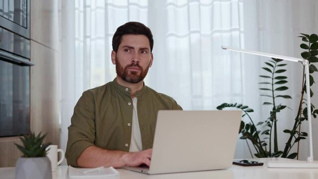 Focused guy with beard and moustache using professional laptop and thoughtfully looking aside. Concentrated distance worker doubting for while and continuing developing plan for project success.
