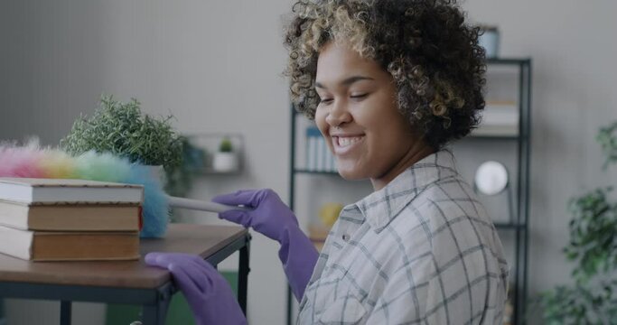 Joyful African American woman dusting shelf with colorful duster wiping furniture smiling focused on clean-up at home. Housework and people concept.