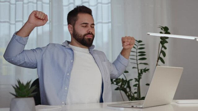 Productive brunette man making break for stretching and warming up during working day by desktop with laptop. Experienced remote programmer relaxing muscles while getting tired from sitting at home.